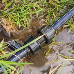 A broken irrigation pipe connector in muddy grass. Water is leaking from the connector and forming a puddle around it.