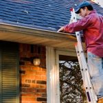 A man wearing a red shirt and a blue hat standing on an aluminum ladder cleaning the gutters on the side of a home.